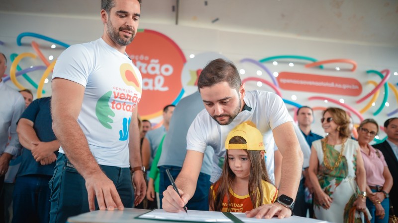 Dois homens vestidos com camisa branca escrita Operação Verão em cores coloridas. Estão assinando um papel com uma criança junto ao meio da foto. Pessoas estão atrás assistindo a assinatura do documento. 