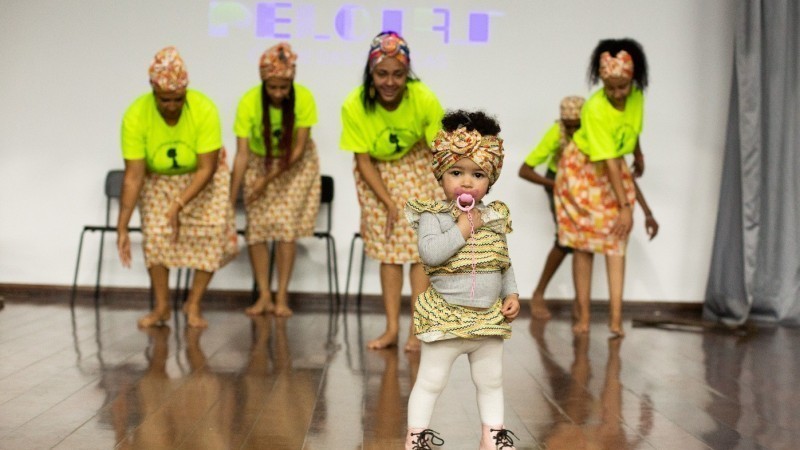 Cinco mulheres e uma criança em um local fechado. As mulheres usam roupas que remetem a culturas africanas e a menina também, usando meia calça branca. As mulheres usam uma blusa amarelo neon. 