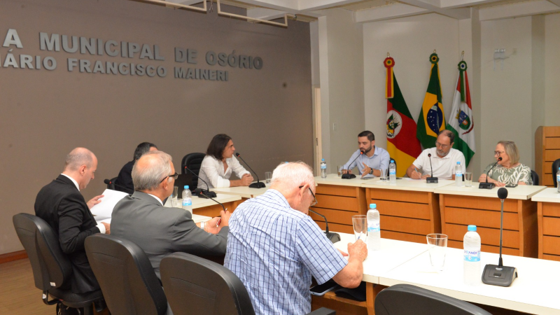 Pessoas reunidas em uma mesa. Algumas estão atrás das bandeiras do Brasil, RS e Osório. 