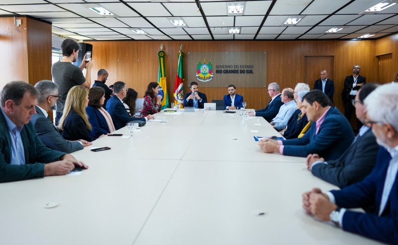 Pessoas sentadas em uma extensa mesa branca. As pessoas, homens brancos usando roupas sociais e mulheres com roupas coloridas e em cores neuras estão virados para a ponta da mesa, onde sentam o governador Eduardo Leite, homem branco usando paletó escuro e o vice-governador Gabriel Souza, usando vestimentas similares. Atrás destes, está a bandeira do Rio Grande do Sul e a do Brasil. A sala tem paredes de madeira e teto branco e preto.