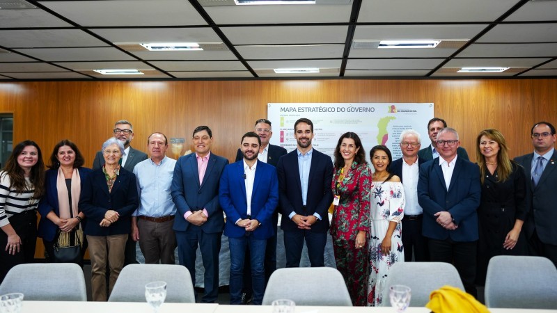 Homens e mulheres posando para uma foto um ao lado do outro, atrás de uma mesa branca com cadeiras cinzas. Centralizados na foto, estão o governador do Rio Grande do Sul, Eduardo Leite, e o vice-governador, Gabriel Souza. As paredes da sala são de madeira e o teto branco e preto.