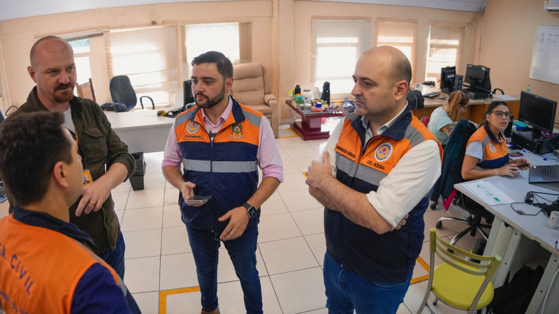 Gabriel conversando com outros três homens na instauração do gabinete de crise avançado da região Metropolitana. è uma sala grande onde outras pessoas estão trabalhando.