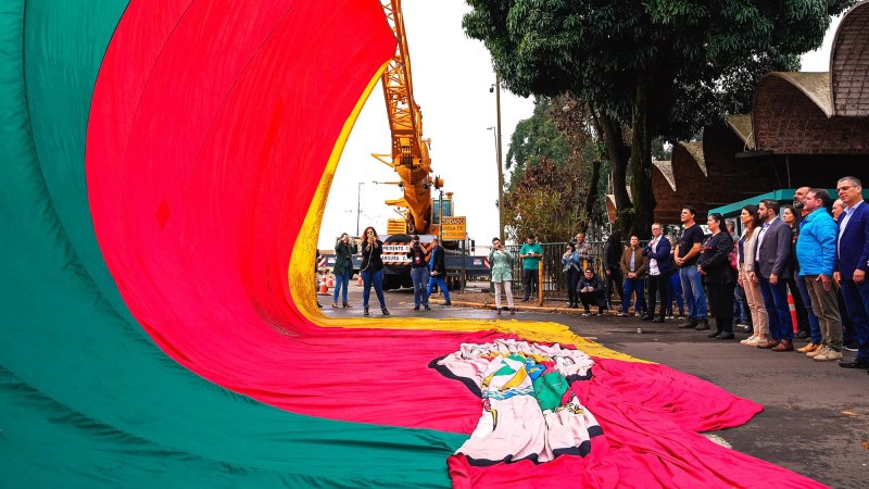 Varia pessoas perfiladas na frente da Ceasa/RS enquanto assistem Ao levantamento de uma bandeira gigantesca do Rio Grande do Sul que fica suspensa por um guincho.