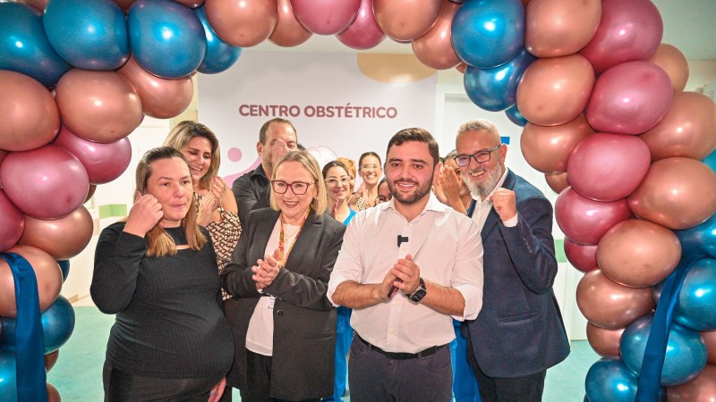 Foto posada. Gabriel, Arita e uma grávida em pé debaixo de um arco de balões aplaudindo a reabertura do centro obstétrico. Há outras pessoas atrás deles participando do momento.