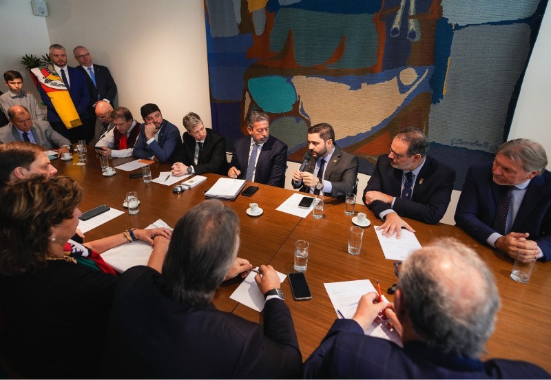 Gabriel falando ao microfone ao lado de Lira sentados à mesa de reunião. A mesa é grande e está tomada por outras pessoas que assistem à reunião.