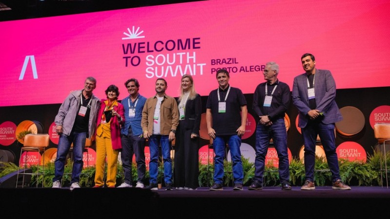 Foto posada frontal do palco do South Summit, em que oito pessoas, dentre elas Gabriel Souza, aparecem de pé, sorrindo. Ao fundo, está um grande painel cor-de-rosa em que está escrito Welcome to South Summit.