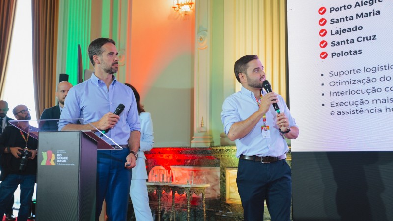 Governador Eduardo Leite ao lado do Vice-governador Gabriel Souza, que está falando ao microfone para a plateia do lado de telão que passa apresentação.