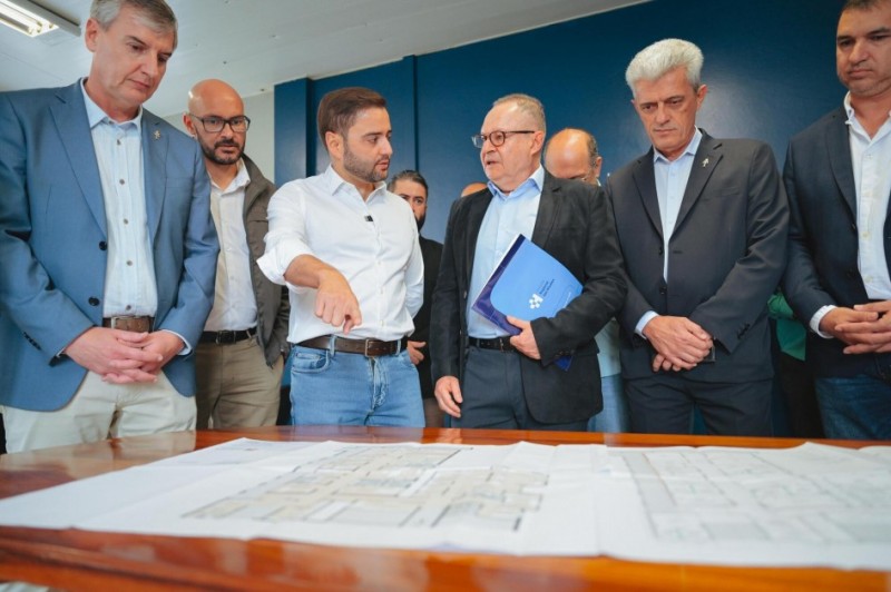 Gabriel aparece em pé, no meio de um grupo de mais cinco homens, conversando com um deles. Todos estão de roupas formais. Gabriel olha de lado aponta com o dedo para um mapa disposto sobre uma mesa.