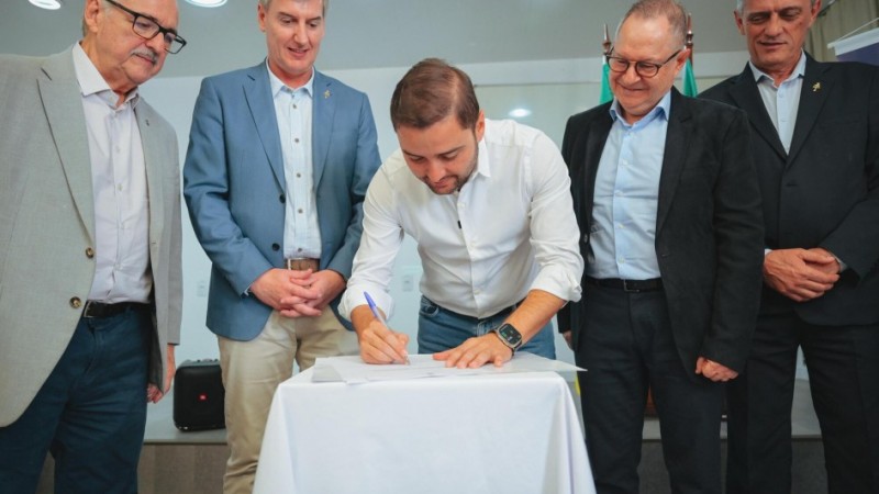 Gabriel aparece no meio da foto, entre quatro homens de roupas formais. Ele está encurvado de pé junto a uma mesa, na qual assina um papel