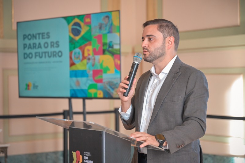 Vice-governador Gabriel Souza falando atrás do púlpito. Uma tela com uma apresentação está atrás dele.