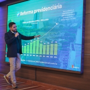 O vice-governador Gabriel Souza está de pé, segurando um microfone, na frente de um painel com um gráfico sobre a reforma previdenciária. Ele aponta para o painel.