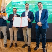 Quatro homens posam para foto em um salão nobre, em frente a um painel digital sobre o Sistema Estadual de Proteção e Defesa Civil. Ao centro, o governador Eduardo Leite segura e exibe um documento oficial aberto, acompanhado pelo vice-governador Gabriel Souza, que aparece imediatamente à esquerda dele (na foto). Os outros dois integrantes, aplaudem.