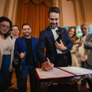 Em um sal&atilde;o com decora&ccedil;&atilde;o formal e cortinas douradas, o governador Eduardo Leite assina um documento sobre uma pequena mesa de madeira enquanto segura um filhotinho de cachorro no colo. Ao lado dele est&atilde;o o vice-governador Gabriel Souza e a secret&aacute;ria do Meio Ambiente, Marjorie Kauffmann. Ao redor, outras pessoas observam a cena. Uma delas tamb&eacute;m tem um c&atilde;o de pequeno porte nos bra&ccedil;os. Ao fundo, h&aacute; um painel institucional parcialmente vis&iacute;vel. A imagem registra um momento formal com a presen&ccedil;a de animais.