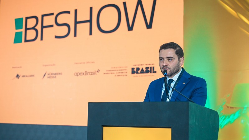Vice-Governador Gabriel Souza fala ao microfone em um p&uacute;lpito durante um evento, vestindo terno e crach&aacute;. Ao fundo, um painel laranja exibe &ldquo;BFSHOW&rdquo; e logotipos institucionais, com ilumina&ccedil;&atilde;o profissional e luzes verdes laterais.