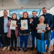 Grupo de oito pessoas posando sob uma tenda em &aacute;rea externa. Algumas seguram documentos com bras&atilde;o e uma pessoa exibe uma chave, sugerindo uma entrega oficial. Ao fundo, h&aacute; uma mesa com toalha azul e branca e um painel com imagem de empreendimento.