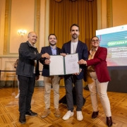 Quatro pessoas, entre elas o governador Eduardo Leite e o vice-governador Gabriel Souza, posam no palco de um sal&atilde;o institucional, segurando um documento aberto durante um ato de assinatura oficial; ao fundo, h&aacute; um tel&atilde;o com texto sobre uma portaria ambiental e, &agrave; esquerda, uma int&eacute;rprete de Libras.