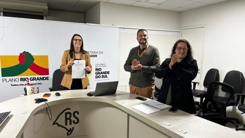 Imagem de uma sala de reuni&atilde;o institucional. Tr&ecirc;s pessoas est&atilde;o atr&aacute;s de uma mesa: uma segura um documento e as outras fazem gesto de aplauso. Sobre a mesa h&aacute; um notebook e objetos pequenos. Ao fundo, aparecem banners com os textos &ldquo;Plano Rio Grande&rdquo; e &ldquo;Secretaria da Sa&uacute;de do Rio Grande do Sul&rdquo;. &Agrave; direita, h&aacute; cadeiras de escrit&oacute;rio.