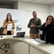 Imagem de uma sala de reuni&atilde;o institucional. Tr&ecirc;s pessoas est&atilde;o atr&aacute;s de uma mesa: uma segura um documento e as outras fazem gesto de aplauso. Sobre a mesa h&aacute; um notebook e objetos pequenos. Ao fundo, aparecem banners com os textos &ldquo;Plano Rio Grande&rdquo; e &ldquo;Secretaria da Sa&uacute;de do Rio Grande do Sul&rdquo;. &Agrave; direita, h&aacute; cadeiras de escrit&oacute;rio.