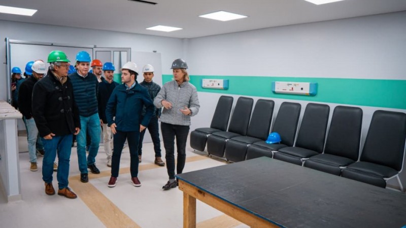 Ambiente interno hospitalar onde um grupo usando capacetes de seguran&ccedil;a percorre o espa&ccedil;o, que possui fileira de cadeiras, equipamentos nas paredes e ilumina&ccedil;&atilde;o forte.

