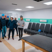 Ambiente interno hospitalar onde um grupo usando capacetes de seguran&ccedil;a percorre o espa&ccedil;o, que possui fileira de cadeiras, equipamentos nas paredes e ilumina&ccedil;&atilde;o forte.

